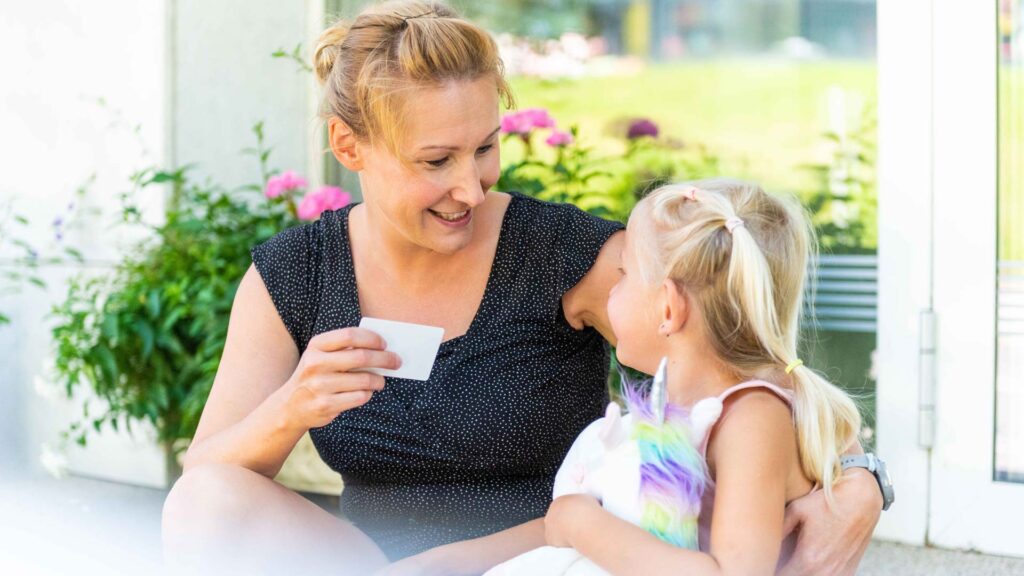 Frau zeigt ihrem Kind die Bürgercard von asut computer, beide sitzen in einer schönen Umgebung mit Blumen, die Karte bietet Gemeindebürgern Vorteile wie vergünstigten Zugang zum Schwimmbad.