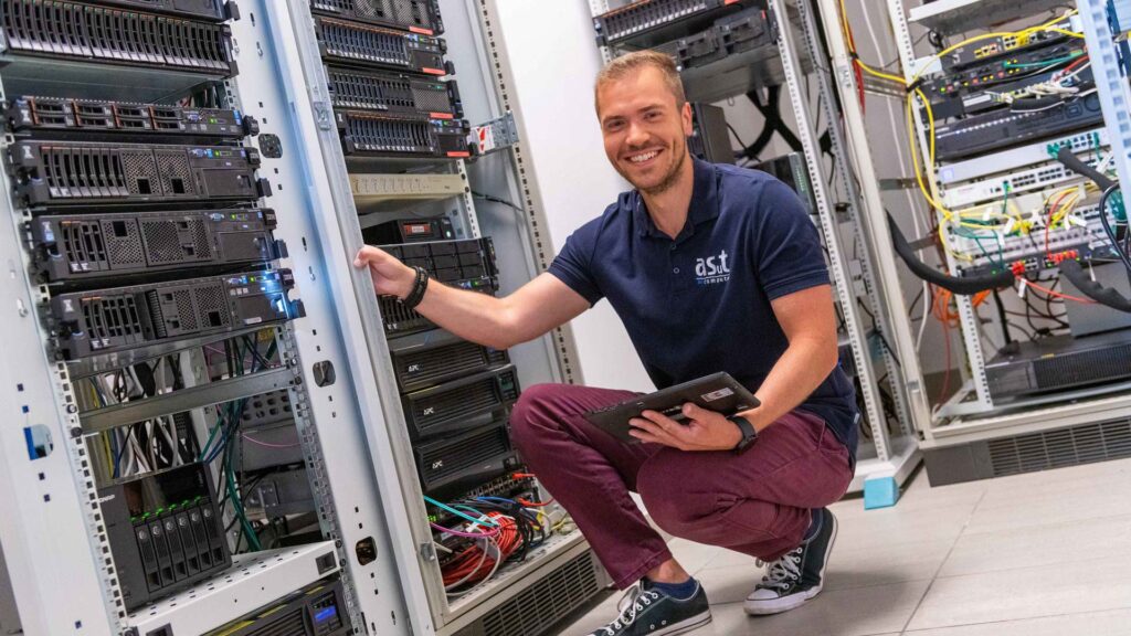 Mann kniet mit Tablet vor einem Serverschrank von asut computer, trägt asut Polo-Shirt und lächelt in die Kamera.