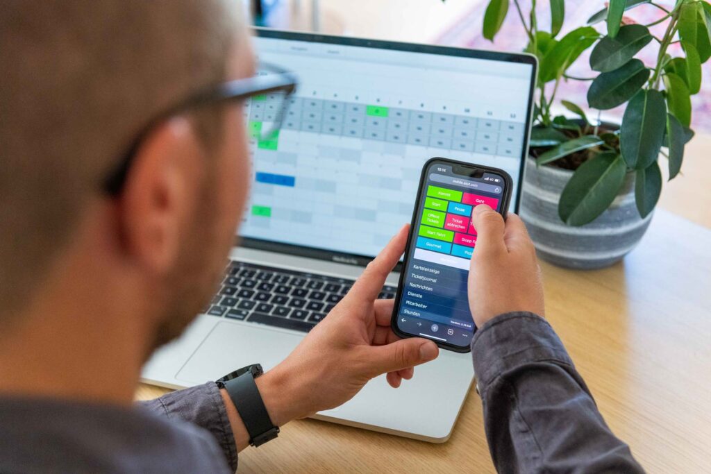 Mann mit Brille hält ein Handy und nutzt Timework von asut computer, Laptop mit geöffneter Zeiterfassung im Hintergrund, Blume auf dem Tisch.