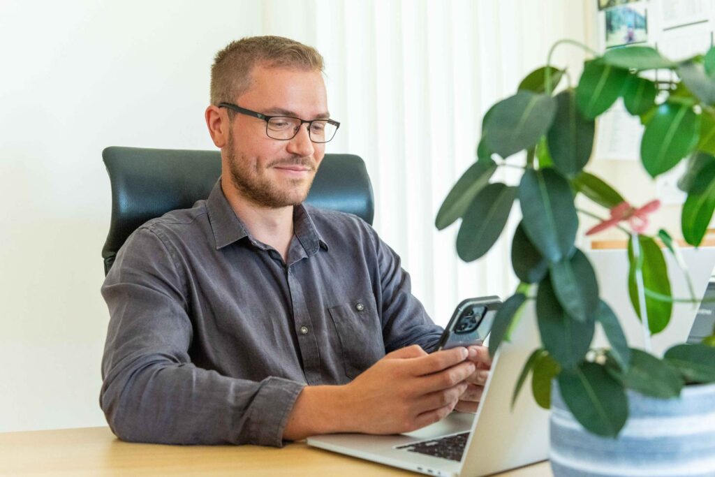 Mann schaut auf sein Handy, sitzt an einem Bürotisch mit Laptop und Blume, nutzt Timework von asut computer.