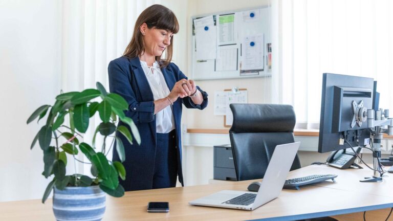 Frau steht vor einem Bürotisch mit Laptop, Handy und Monitor, schaut auf ihre Uhr.