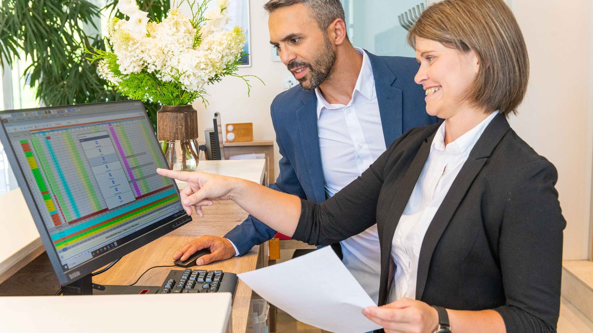 Frau mit Zettel zeigt einem Mann auf den Monitor die Hotelsoftware von asut computer, Blume und Hotellobby im Hintergrund.