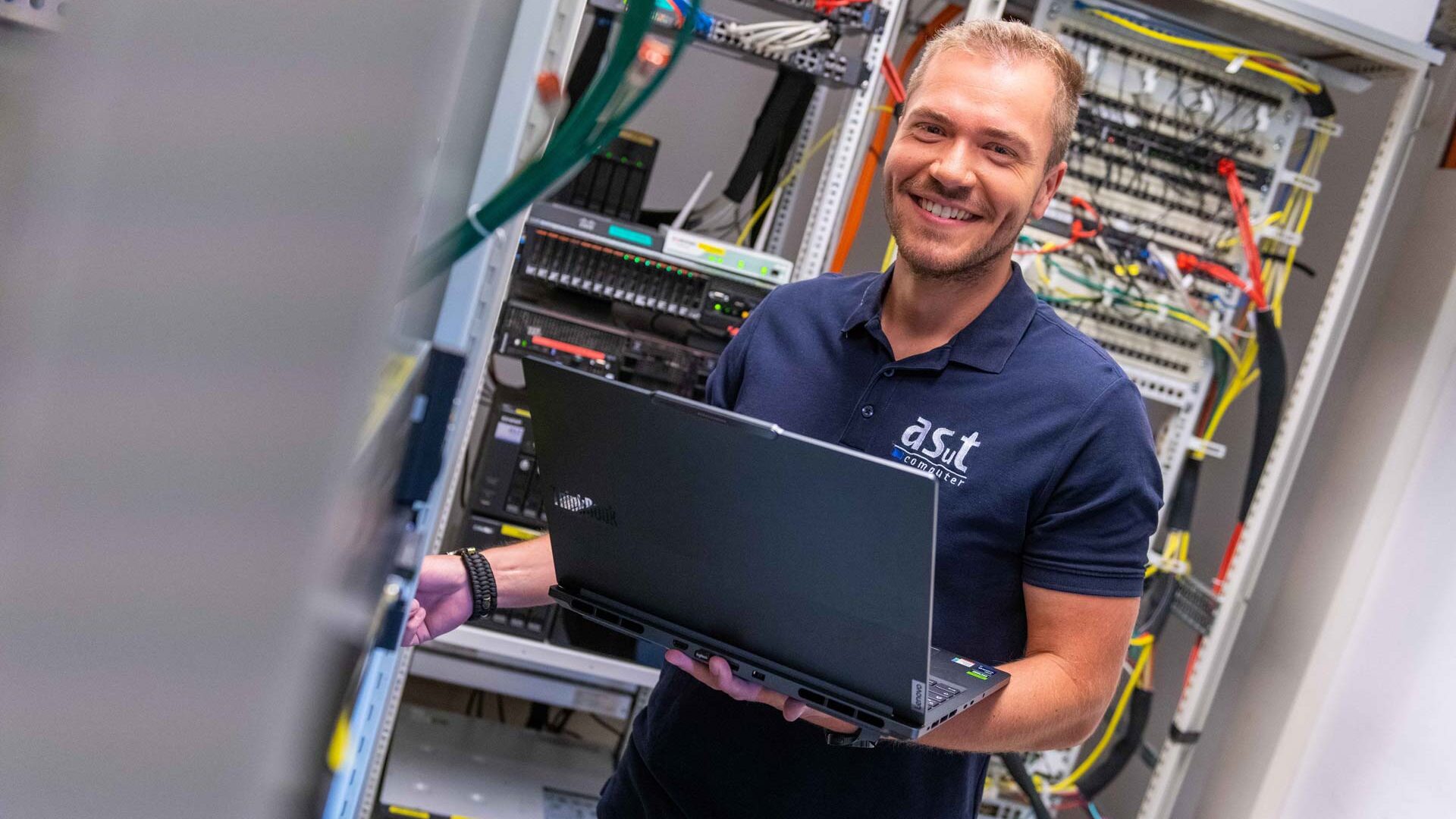 Mann mit Laptop im Serverraum von asut computer, lächelt in die Kamera und trägt asut computer Polo-Shirt.